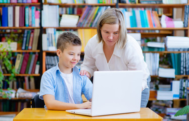 A teacher or tutor helps a boy with a blue shirt sitting at a laptop in a library study. Learning assistance, concept support