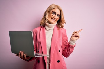 Middle age beautiful blonde business woman working using laptop over pink background very happy pointing with hand and finger to the side