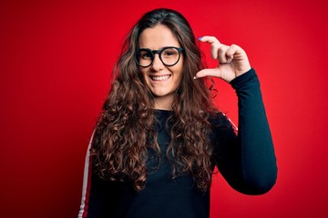 Young beautiful woman with curly hair wearing sweater and glasses over red background smiling and confident gesturing with hand doing small size sign with fingers looking and the camera. Measure