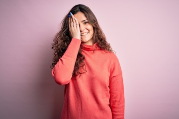 Young beautiful woman with curly hair wearing turtleneck sweater over pink background covering one eye with hand, confident smile on face and surprise emotion.