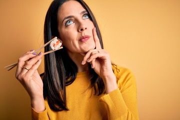 Young brunette woman with blue eyes eating salmon maki sushi using chopsticks serious face thinking...