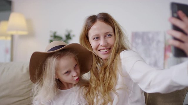Medium shot of adorable little girl in wide brim hat and her happy mother sitting on couch and taking selfie on mobile phone