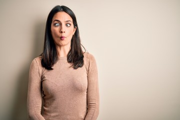 Obraz premium Young brunette woman with blue eyes wearing casual sweater over isolated white background making fish face with lips, crazy and comical gesture. Funny expression.