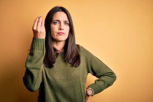 Young brunette woman with blue eyes wearing green casual sweater over yellow background Doing Italian gesture with hand and fingers confident expression