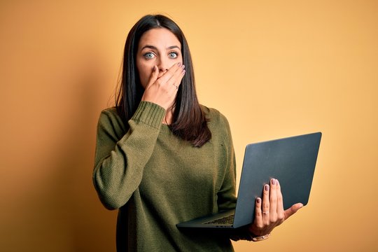 Young brunette woman with blue eyes working using computer laptop over yellow background shocked covering mouth with hands for mistake. Secret concept.