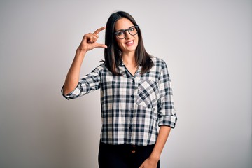 Young brunette woman with blue eyes wearing casual shirt and glasses over white background smiling and confident gesturing with hand doing small size sign with fingers looking and the camera. Measure