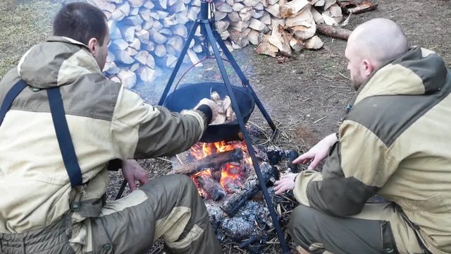 Two men in military clothing Cooking outdoors in cast-iron cauldron. Cooking on a fire. Food in a cauldron on a fire in nature