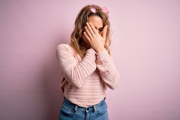 Young beautiful blonde woman wearing casual sweater and sunglasses over pink background with sad expression covering face with hands while crying. Depression concept.
