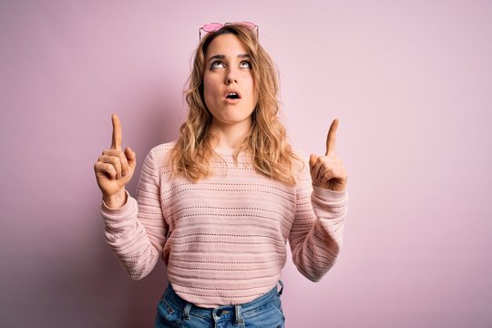 Young beautiful blonde woman wearing casual sweater and sunglasses over pink background amazed and surprised looking up and pointing with fingers and raised arms.