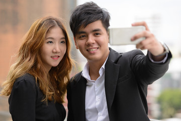 Happy young Asian business couple taking selfie together in the city