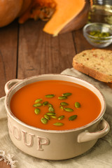 Bowl with pumpkin soup served with seeds on sackcloth with ingredients on wooden background. Carrot organic vegetarian bread, seeds and slice of ripe pumpkin. Menu, recipe