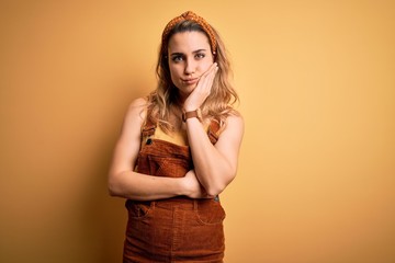 Young beautiful blonde woman wearing overalls and diadem standing over yellow background thinking looking tired and bored with depression problems with crossed arms.