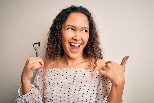 Young beautiful woman with curly hair holding eyelases curler over white background pointing and showing with thumb up to the side with happy face smiling