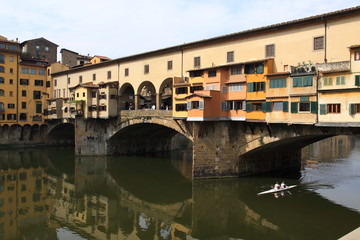Fototapeta na wymiar a boat with two rowers runs under a ponte vecchio bridge in florence on the arno river