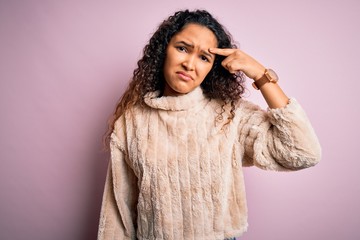 Young beautiful woman with curly hair wearing casual sweater standing over pink background pointing unhappy to pimple on forehead, ugly infection of blackhead. Acne and skin problem