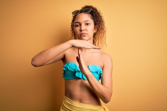 Young beautiful american woman on vacation wearing bikini over isolated yellow background Doing time out gesture with hands, frustrated and serious face