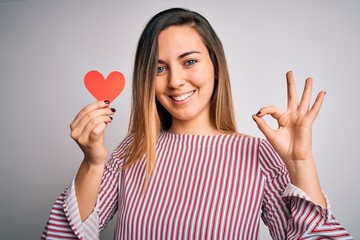 Young beautiful blonde romantic woman holding red paper heart over white background doing ok sign...