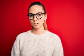 Beautiful blonde woman with blue eyes wearing sweater and glasses over red background with serious expression on face. Simple and natural looking at the camera.
