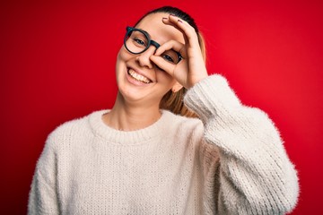 Beautiful blonde woman with blue eyes wearing sweater and glasses over red background doing ok gesture with hand smiling, eye looking through fingers with happy face.