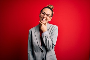 Young beautiful blonde businesswoman with blue eyes wearing glasses and jacket looking confident at the camera smiling with crossed arms and hand raised on chin. Thinking positive.