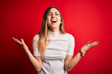Beautiful blonde woman with blue eyes wearing casual white t-shirt over red background crazy and mad shouting and yelling with aggressive expression and arms raised. Frustration concept.