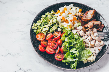 American traditional Cobb salad with chicken, avocado, tomatoes, eggs, bacon and cheese in a black plate on a marble table.