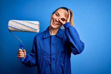 Young beautiful blonde painter woman with blue eyes painting wearing uniform using roller with...