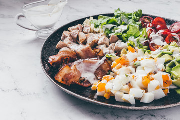 American traditional Cobb salad with chicken, avocado, tomatoes, eggs, bacon and cheese in a black plate on a marble table.