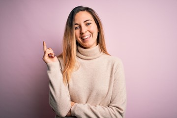 Beautiful blonde woman with blue eyes wearing turtleneck sweater over pink background with a big smile on face, pointing with hand and finger to the side looking at the camera.
