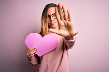 Young beautiful blonde romantic woman wearing glasses holding big pink heart with open hand doing stop sign with serious and confident expression, defense gesture