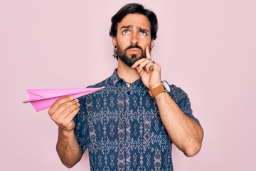 Young handsome hispanic bohemian hipster man with bear holding paper plane serious face thinking...