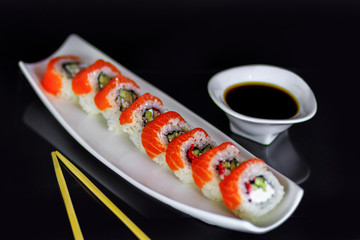 Sushi roll with red salmon fish, cucamber, and white cheese on a plate with say sauce and chopsticks on black background.