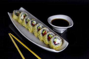 Sushi roll with salmon fish, cucamber, and white cheese on a plate with say sauce and chopsticks on black background.
