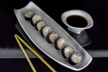 Sushi roll with salmon fish, cucamber, and white cheese on a plate with say sauce and chopsticks on black background.