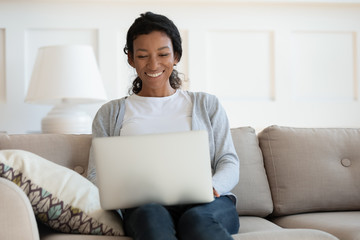 Smiling millennial happy african american woman sit on sofa, typing, hold computer on her lap and look at laptop screen. Middle aged woman working at home, online studying or shopping - Powered by Adobe