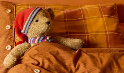 Cute, cuddly plush bear with woollen scarf & bed cap, tucked up in bed, with red and orange plaid pillow, with decorative buttons and ribbing. Interior landscape image with space for copy. England.