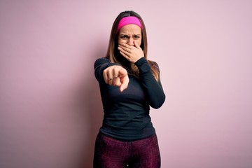 Young blonde fitness woman wearing sport workout clothes over isolated background laughing at you, pointing finger to the camera with hand over mouth, shame expression