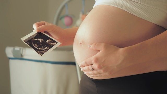 Pregnant woman middle body section holding the echography photograph of her baby in front of her belly.