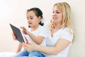 Happy family mom and kid daughter using digital tablet sitting on sofa, smiling parent mother with child girl holding tablet looking at screen do online shopping make video call watch cartoons