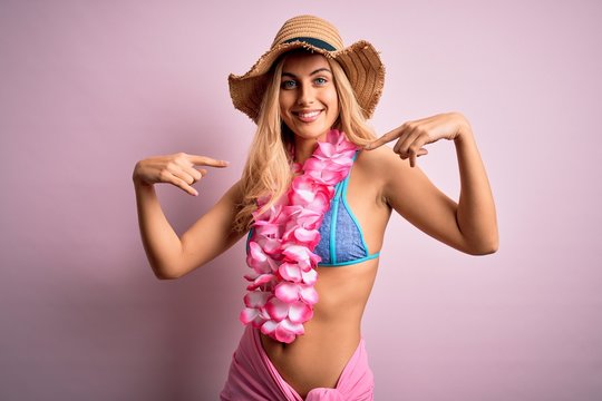 Young beautiful blonde woman on vacation wearing bikini and hat with hawaiian lei flowers looking confident with smile on face, pointing oneself with fingers proud and happy.