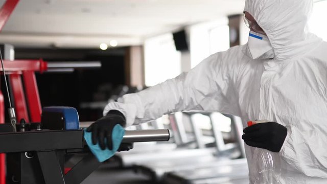 Hazmat worker disinfects gym fitness equipment from coronavirus covid-19 hazard with antibacterial sanitizer sprayer and wipe. Man in protective suit cleans training apparatus at workout area.