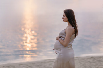 Fototapeta na wymiar Beautiful pregnant woman on the coast sunset. The concept of expectation of a child calm pacification silence. Gentle tones of tenderness. Place for text. Young mom. Easy pregnancy relax new life