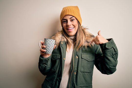 Young beautiful brunette woman wearing snow clothes drinking mug of coffee with surprise face pointing finger to himself