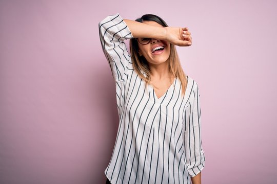 Young beautiful woman wearing casual striped t-shirt and glasses over pink background covering eyes with arm smiling cheerful and funny. Blind concept.