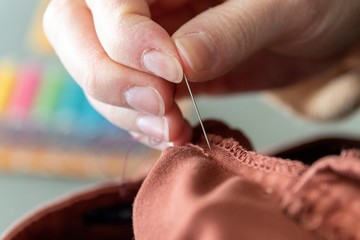 A close up portrait of a hand of someone holding a needle, sewing a hole in a pair of pants at the seam. The needle is about to be put through the fabric to repair it with a thread.