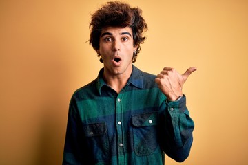 Young handsome man wearing casual shirt standing over isolated yellow background Surprised pointing...