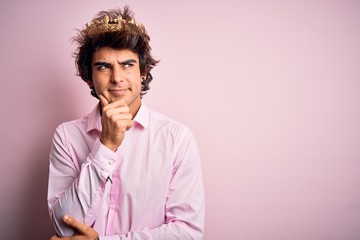 Fototapeta na wymiar Young handsome man wearing king crown standing over isolated pink background with hand on chin thinking about question, pensive expression. Smiling with thoughtful face. Doubt concept.