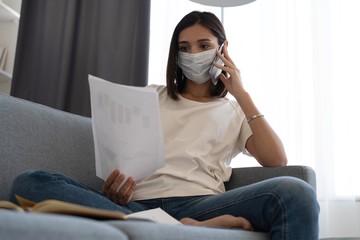 Young woman wearing protective mask in quarantine and using smartphone while sitting on comfortable sofa