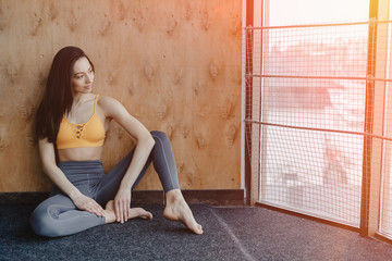 Young attractive fitness girl sitting on the floor near the window on the background of a wooden wall, resting on yoga classes