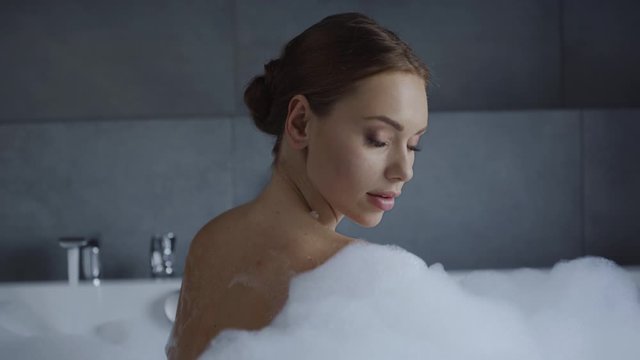 beautiful young woman washing shoulder and looking at camera in bubble bath